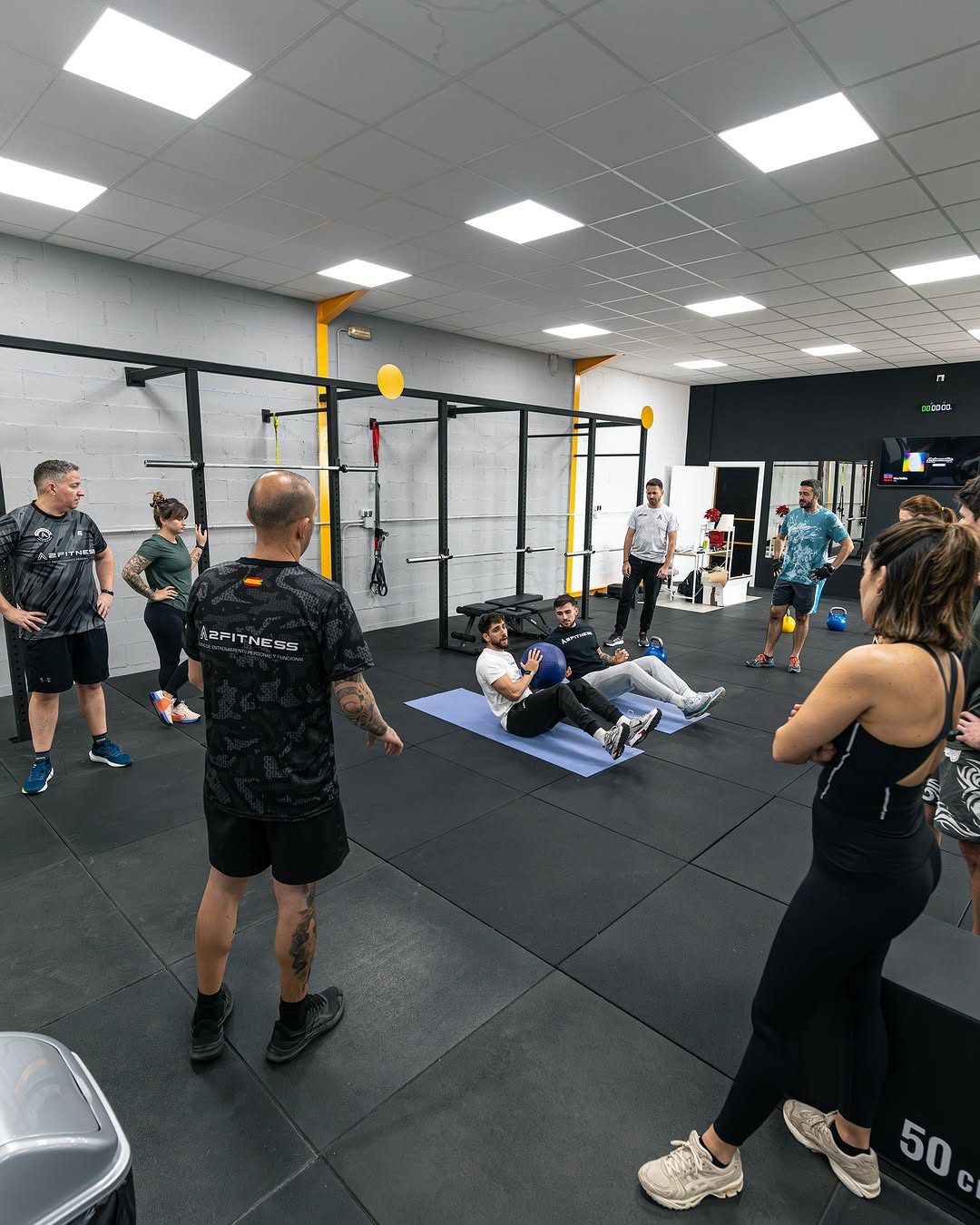 Grupo de personas participando en una sesión de entrenamiento en un gimnasio moderno, con un instructor guiando ejercicios de acondicionamiento físico. Se observan diferentes equipos de entrenamiento y un ambiente motivador.